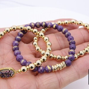 May include: Three gold beaded bracelets with purple and gold beads. The bracelet on the bottom has a gold charm with a purple stone.