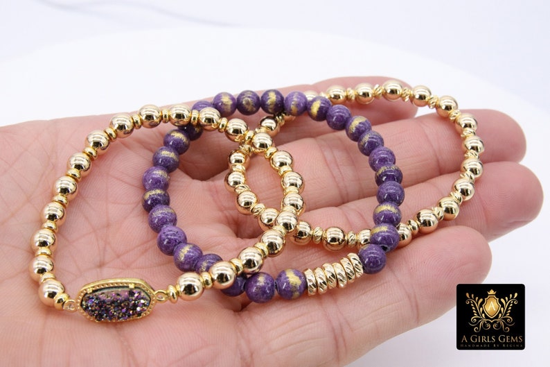 May include: Three gold beaded bracelets with purple and gold beads. The bracelet on the bottom has a gold charm with a purple stone.