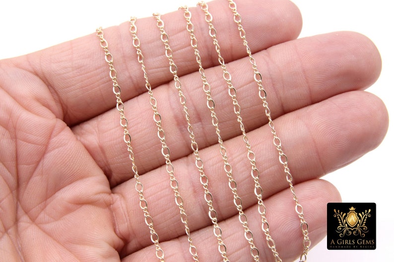 May include: A close-up of a gold chain necklace. The chain is made of small, oval-shaped links. The chain is lying on a hand, and the background is white.