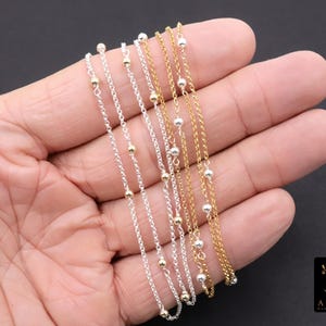 Könnte beinhalten: Mehrere zarte Halsketten mit kleinen Perlen und Kettengliedern in Silber- und Goldtönen werden gezeigt. Die Halsketten werden in einer Hand vor dunklem Hintergrund gehalten. Die Halsketten haben ein schlichtes, elegantes Design.