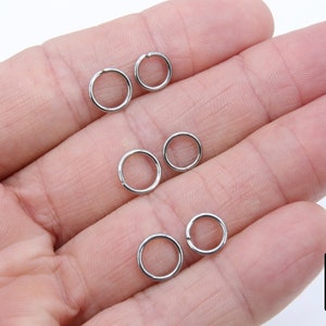 May include: Six silver jump rings, also known as split rings, are shown on a hand. These rings are used for attaching charms, pendants, or other jewellery components.
