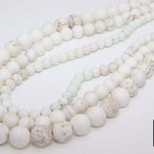 May include: A strand of white and cream colored round beads with a speckled pattern. The beads are strung on a white cord and are arranged in a circular pattern.