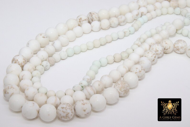 May include: A strand of white and cream colored round beads with a speckled pattern. The beads are strung on a white cord and are arranged in a circular pattern.