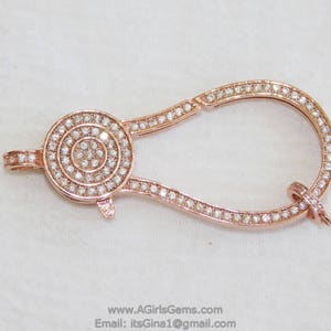 May include: A rose gold-toned clasp with a round, pave-set diamond design. The clasp is shaped like a teardrop and has a hinged opening.