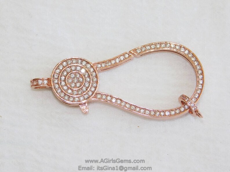May include: A rose gold-toned clasp with a round, pave-set diamond design. The clasp is shaped like a teardrop and has a hinged opening.