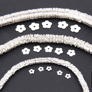 May include: Silver metal beads in various sizes, arranged in a circular pattern. The beads are flat and have a slightly wavy texture. There are also several small silver flower-shaped charms scattered around the beads.
