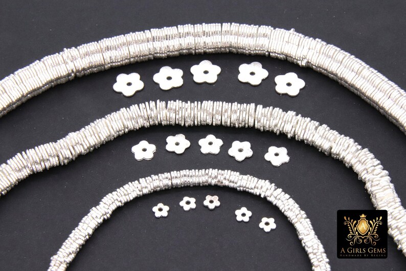 May include: Silver metal beads in various sizes, arranged in a circular pattern. The beads are flat and have a slightly wavy texture. There are also several small silver flower-shaped charms scattered around the beads.