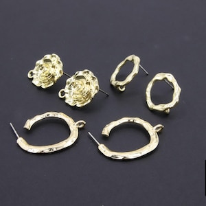 May include: Gold-toned metal earrings in various styles, including floral studs, irregular circle studs, and oval hoop earrings. The earrings are displayed on a dark gray surface. The image also includes the logo of "A Girls Gems".