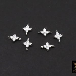 May include: Six small, silver-toned flower-shaped charms with clear, sparkling crystals. Each charm has four petals and a loop for attaching to jewellery. The charms are displayed on a dark background.