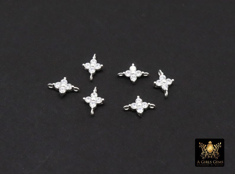 May include: Six small, silver-toned flower-shaped charms with clear, sparkling crystals. Each charm has four petals and a loop for attaching to jewellery. The charms are displayed on a dark background.