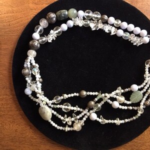 May include: A multi-strand necklace with white, green, and silver beads. The necklace is made of various materials, including glass, metal, and stone. The necklace is displayed on a black velvet surface.