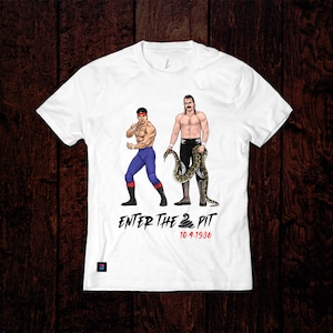 以下が含まれることがあります： 2人のレスラーが描かれた白いTシャツ。一人はヘビを持っています。レスラーの下には「Enter the 2 Pit 10-9-1996」という文字が印刷されています。