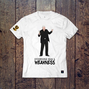 May include: White t-shirt featuring a cartoon illustration of a man holding a glass of champagne and making a hand gesture. The text "EVERYONE HAS A WEAKNESS" is printed below the illustration.