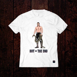 以下が含まれることがあります： 白いTシャツ。口ひげを生やした男性がヘビを持っている漫画のイラスト。テキスト「DDT = THE END」は濃い青色で印刷されています。デザインは漫画のイラストです。