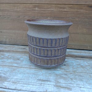 May include: A handcrafted, cylindrical ceramic vase in shades of brown and tan. The vase features a textured, carved design around the middle section. The top rim is slightly flared, adding a touch of elegance to this rustic home decor item.