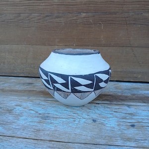May include: A small, round, off-white pottery vessel with a black and white geometric design. The rim is dark, and the base features a series of triangles. The pottery is set against a wooden background.