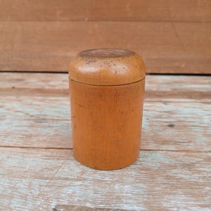 May include: A small, cylindrical wooden container with a rounded lid. The container is a warm, light brown color, with visible wood grain and some wear on the lid. The design is simple and classic, suitable for storage.