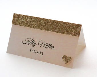 Puede incluir: Una tarjeta de mesa de purpurina dorada con fondo rosa claro. La tarjeta tiene el texto "Kelly Miller" y "Mesa 15" impreso en negro. Un pequeño corazón de purpurina dorada se encuentra en la esquina inferior derecha.