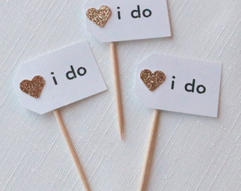 Pode incluir: Três etiquetas de cartão branco com corações de glitter dourado e o texto "i do" em cada uma. As etiquetas estão presas a espetos de madeira.