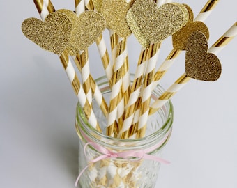 Könnte beinhalten: Goldene Glitzer-Herz-Papierstrohhalme mit weißen und goldenen Streifen in einem durchsichtigen Glas mit einem rosa Band.