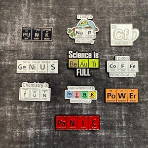 May include: A collection of nine enamel pins featuring periodic table elements arranged to spell words including PUNK, NAP, Cf, GENIUS, BEAUTIFUL, COFFEE, FUN, THINK, POWER, and PANIC, with various colours and scientific text designs.