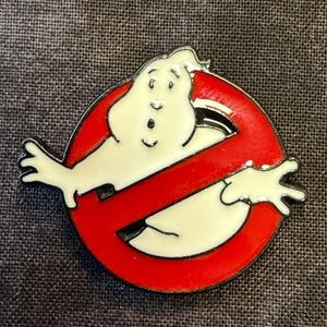 May include: A Ghostbusters logo pin featuring the iconic ghost figure within a red circle with a diagonal red line. The ghost is white with black accents, set against a dark gray background.