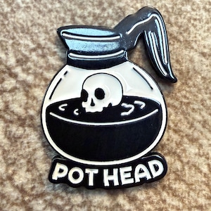 May include: A black and white enamel pin shaped like a coffee pot with a skull submerged in the dark liquid inside. The design features a handle on the right side and a banner at the bottom with the text POT HEAD in bold white capital letters.