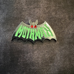 May include: A silver-colored Mothman pin in the shape of a bat. The pin has green lettering that spells out "MOTHMAN" and red eyes. The pin is on a dark background.