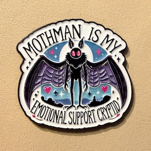 May include: A die-cut sticker featuring a stylized Mothman figure with large wings and glowing red eyes. The text "Mothman is my Emotional Support Cryptid" is displayed in a curved design, with a blue and pink starry background.