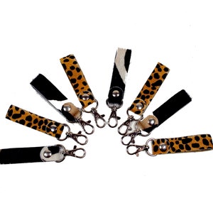 Può includere: Una collezione di portachiavi con vari motivi animalier. I portachiavi presentano un moschettone in metallo e un cinturino in pelle con motivi tra cui stampa leopardata, pelle di mucca bianca e nera e nero tinta unita.