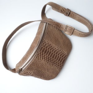 Könnte beinhalten: Eine braune Leder-Bauchtasche mit Schlangenhautmuster und Reißverschluss. Die Tasche hat einen langen verstellbaren Riemen.