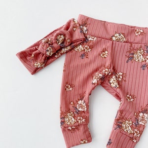 May include: A pink floral print baby leggings set with a matching headband. The leggings have a ribbed texture and a floral pattern in shades of brown, white, and blue.