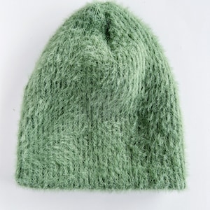 May include: A sage green beanie hat with a fuzzy, textured appearance. The hat is made of a soft, knitted material and is designed to keep the head warm. The hat is a solid color and is suitable for cold weather.