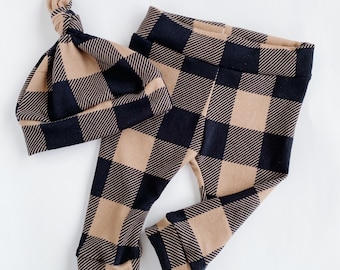 May include: A matching set of baby clothing, including a knotted hat and leggings. The set features a black and tan plaid pattern. The leggings have a fitted design with cuffs at the ankles. The hat is knotted at the top.