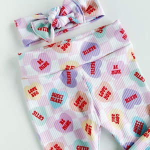 May include: A matching set of baby leggings and a headband. The white ribbed fabric is patterned with colorful candy hearts with phrases like "Be Mine" and "Love Bug" in red. The headband has a knotted bow.