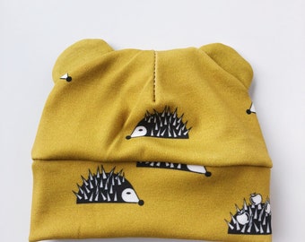 May include: Mustard yellow baby hat with a repeating pattern of black and white hedgehogs. The hat has two small ear-like protrusions at the top. The fabric is soft and the hat is designed for comfort and style.