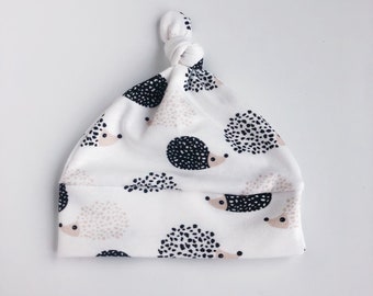 May include: White baby hat with a knot on top. The hat has a pattern of black and beige hedgehogs.