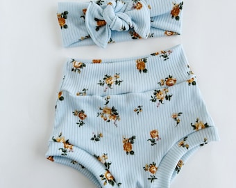 May include: A baby blue ribbed knit headband with a bow and matching high-waisted shorts. Both items feature a floral pattern in yellow and brown. The set is designed for infants.