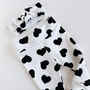 May include: A white baby outfit set featuring black heart patterns. The set includes a pair of leggings and a matching knotted headband. The leggings have a high waistband and fitted cuffs.