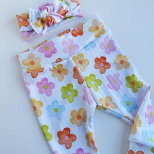 May include: White ribbed leggings and a matching headband, both adorned with a colorful floral pattern. The flowers are in shades of pink, orange, yellow, green, and blue. The headband is knotted at the top.