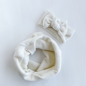 May include: A white ribbed knit headband and matching neck warmer. The headband is tied in a bow.