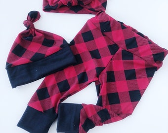 May include: A baby clothing set featuring a red and black buffalo plaid pattern. The set includes a knotted headband, a beanie, and leggings. The cuffs and the bottom of the beanie are solid navy blue.
