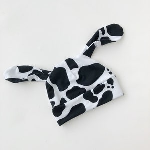 May include: A baby hat with a black and white cow print. The hat has two long, floppy ear-like extensions on top. The hat is made of a soft fabric and is designed to fit a baby's head.