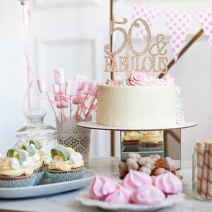 May include: A white cake with pink and blue frosting and a gold '50 & Fabulous' topper. The cake is on a white cake stand with a mirrored base. There are cupcakes with rainbow sprinkles and pink and white meringues on the table.