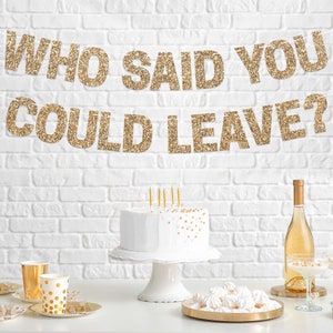 Gold glitter banner with the text "Who Said You Could Leave?" hanging above a white cake with gold sprinkles on a white cake stand. There are gold and white polka dot paper cups and a bottle of white wine on a table with a white brick wall background.
