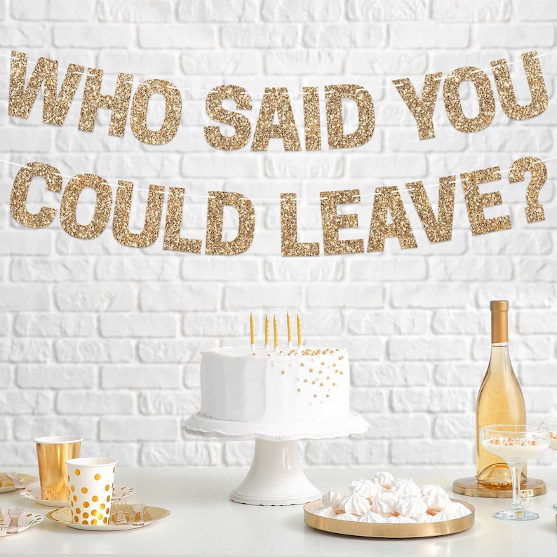 Gold glitter banner with the text "Who Said You Could Leave?" hanging above a white cake with gold sprinkles on a white cake stand. There are gold and white polka dot paper cups and a bottle of white wine on a table with a white brick wall background.