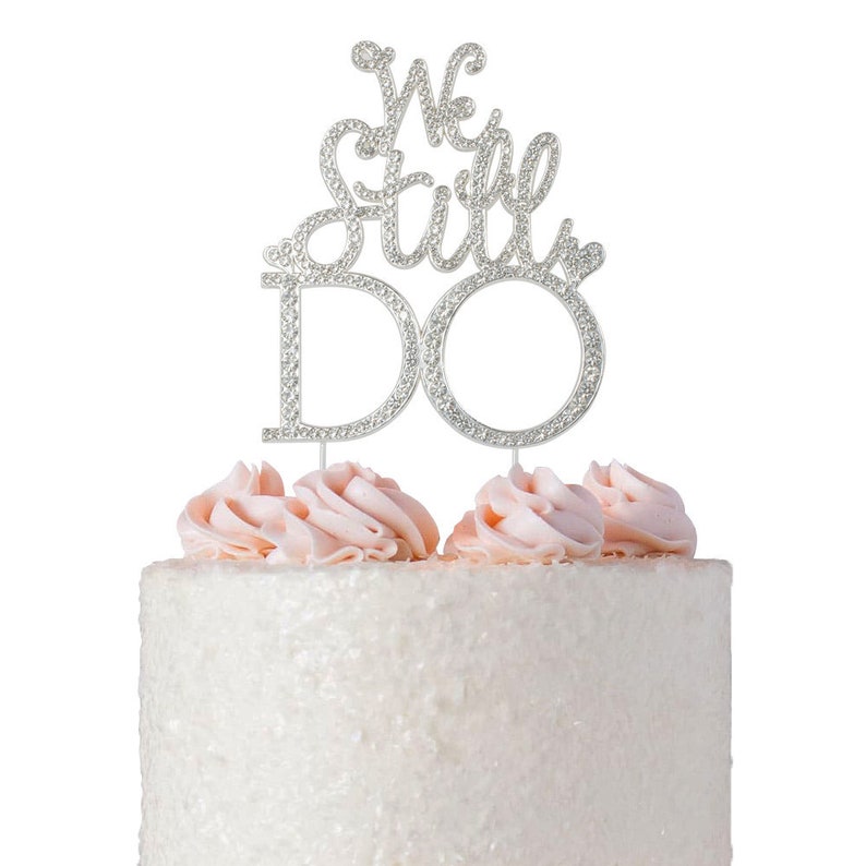 May include: A silver cake topper with the words "We Still Do" encrusted with rhinestones. The topper is placed on a white cake with pink frosting swirls. A celebratory cake decoration for a wedding or anniversary.