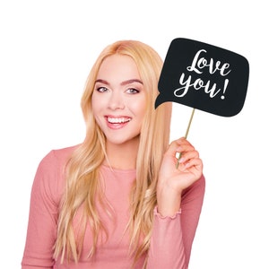 May include: A woman holds a black speech bubble prop with the text "Love you!" written in white. She is smiling and wearing a pink long-sleeved shirt.