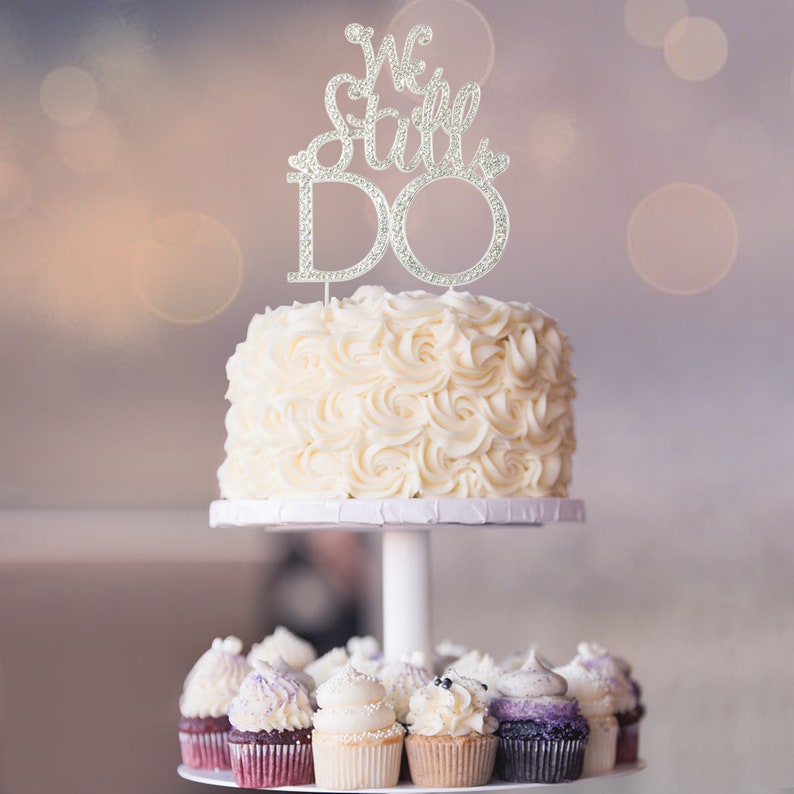 May include: A tiered cake with a white frosting swirl design, topped with a silver cake topper that reads "We Still Do". The bottom tier features cupcakes with various frosting colors and decorations. The cake is on a white stand.