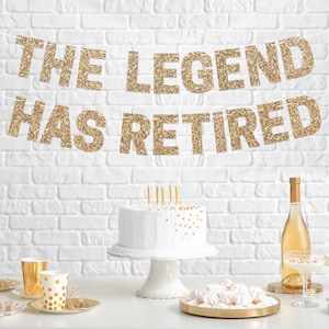 Gold glitter banner with the text "The Legend Has Retired" hanging on a white brick wall. A white cake with gold sprinkles sits on a white cake stand in front of the banner. There are gold and white party supplies on the table.